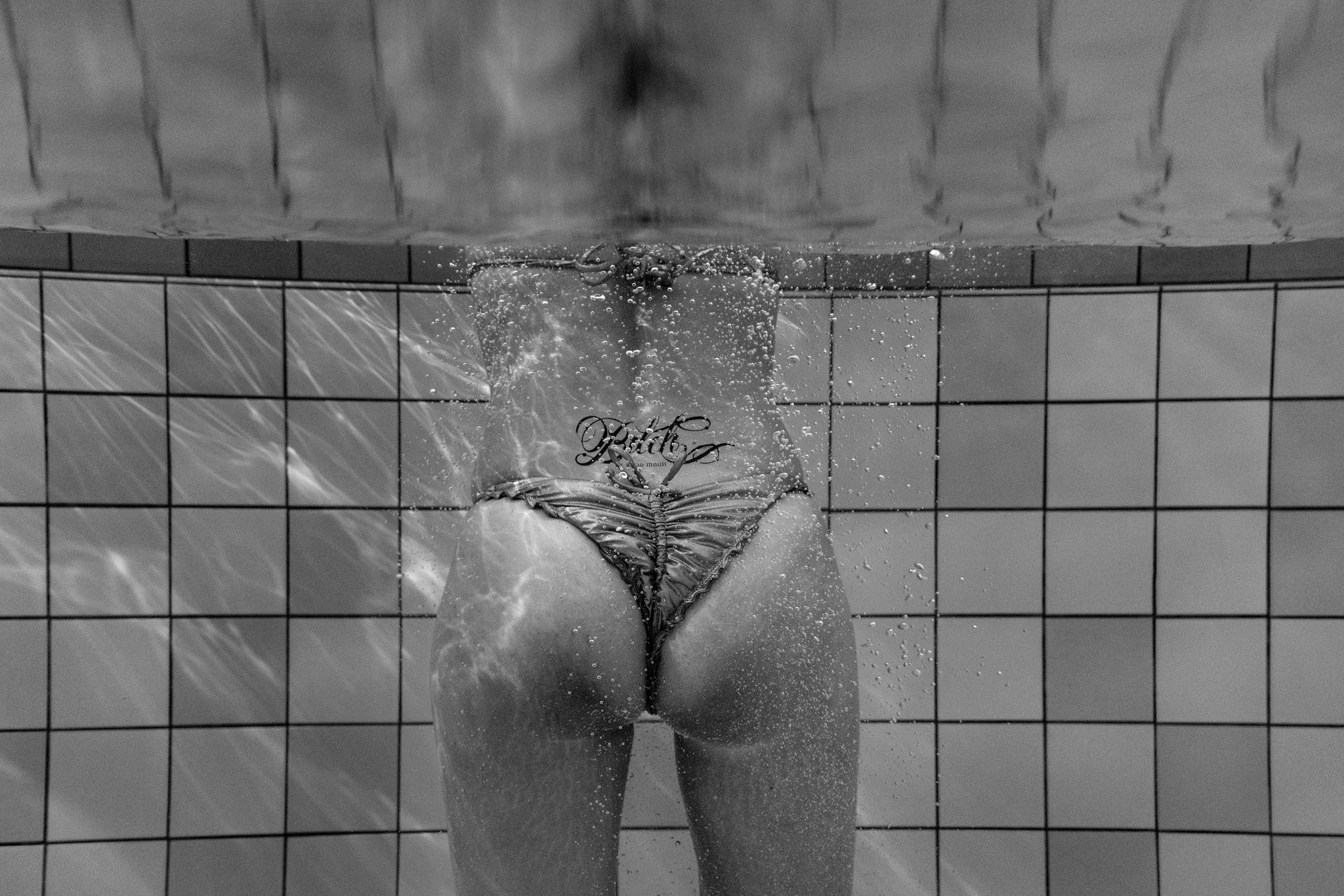Girl in golden bikini with a tramp stamp 'Bitch' on her back underwater. Captured in black & white.